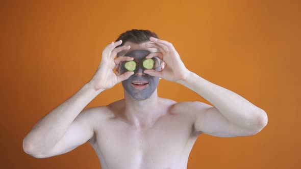 Young Man Uses a Gray Clay Face Mask with Cucumber Slices, Expresses Positive Emotions, Isolated