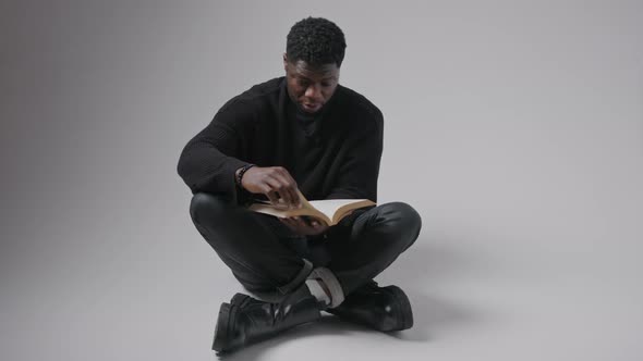 Focused Interested Black Guy Wearing Black Clothes Sitting Crosslegged on the Floor Reading a Book