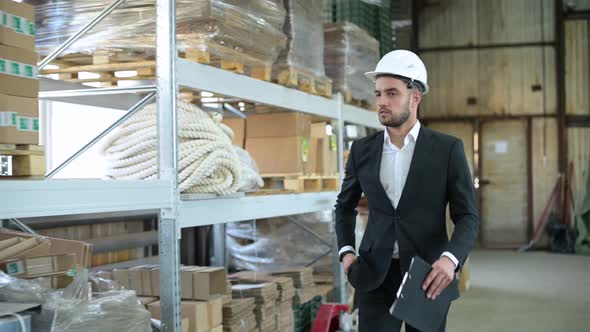 Young boss in a suit and white hard hat walks through a warehouse