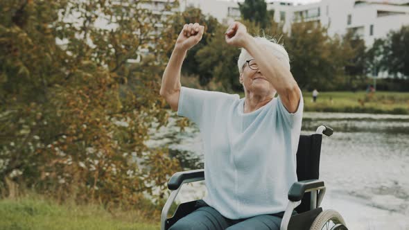 Happy Old Woman, Grandmother, Dancing with Hands in the Wheelchair Near the River 