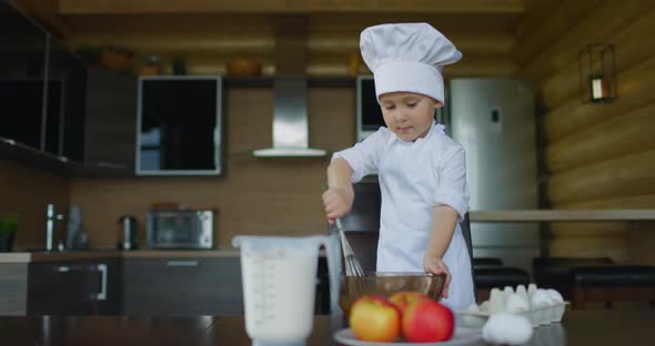 Little Boy in Cook's Costume Kneads Pie Dough with Whisk in the Kitchen
