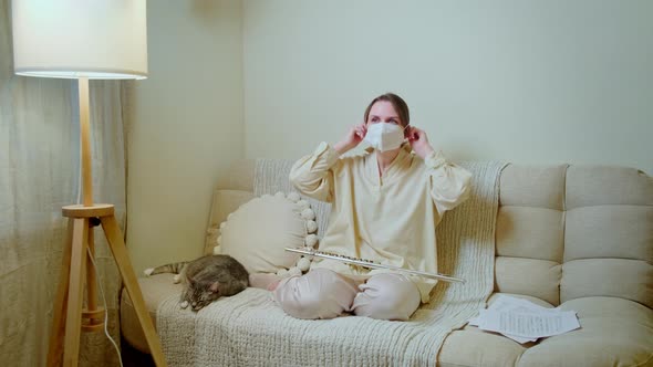 Woman musician with flute in medical face mask playing at home on sofa in living room