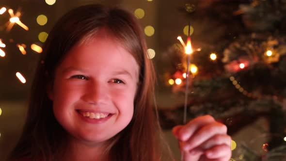 Cute Little Girl Near Christmas Tree Branch with Bengal Lights Against the Background of Sparkling