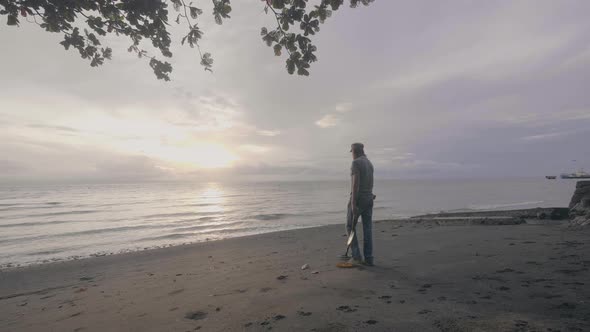 Man Treasure Hunter with a Metal Detector Works on the Ocean Sand Beach at Sunset.