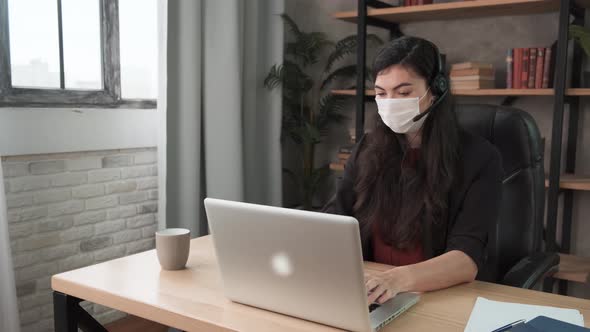 Young Smiling Woman Employee in a Medical Facial Mask Works in the Office at the Computer. Brunette