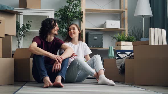 Young Couple Talking and Kissing Sitting on Floor with Boxes Moving in New Flat