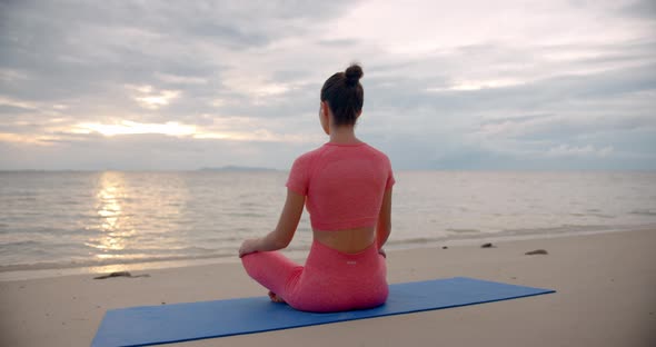 Woman on the Beach Seats on Yoga Mat She is in Lotus Pose and Meditating Against the Sunset