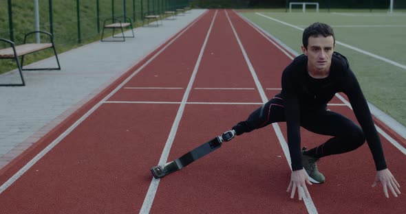 Handsome Disabled Male Sportsman with Prosthetic Running Blades Stretching Limbs at Sports Field