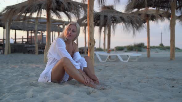 in the Evening at Sunset One Beautiful Woman Sits on the Sand By the Ocean Dressed in a White Shirt