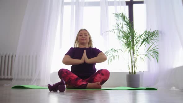 Yoga Indoors  Blonde Overweight Smiling Woman Meditating on Yoga Mat in White Modern Studio