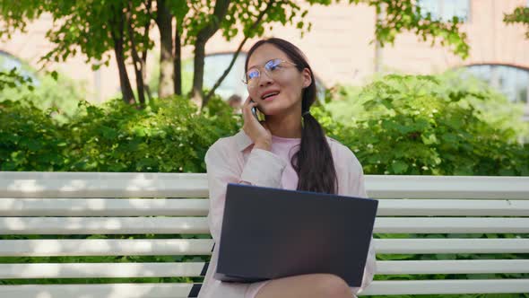 Young Asian Freelance Woman Talking on Phone Working on Laptop Sitting on Bench