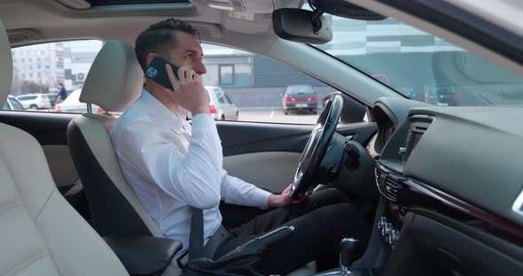 Young Male Businessman is Sitting in the Driver's Seat in the Car and Talking on a Cell Phone