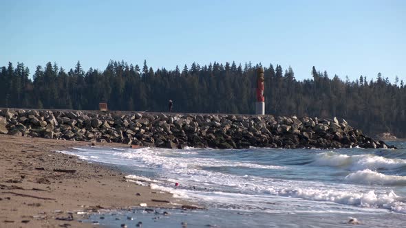 Young woman walking away from a tall statue on a rocky pier on the beach with waves hitting the shor