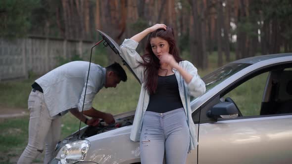 Portrait of Beautiful Sad Woman Standing at Broken Car with Man Repairing Vehicle at Open Hood