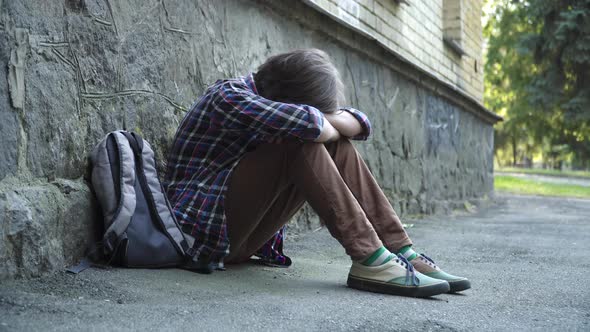 Side View of Crying Bullied Schoolboy Sitting Outdoors at Rock Wall. Wide Shot of Sad Caucasian