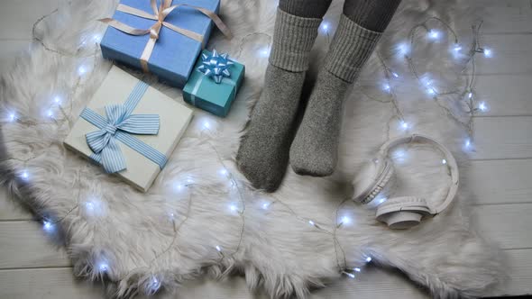 Pair of Female Feet in Gray Socks Next To Blue Gift Boxes, Headphones and a Luminous Garland. Woman