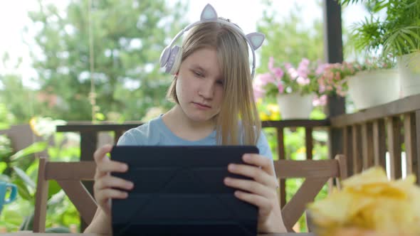 Teenage Girl Sits At Wooden Table In a Summer Cafe In Headphones With Phone