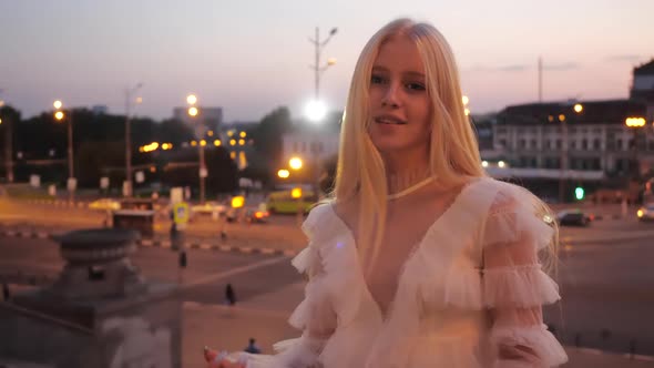 Cute Lovely Young Blonde Girl in White Dress Sips Long Silky Hair and Smiles Against the Background