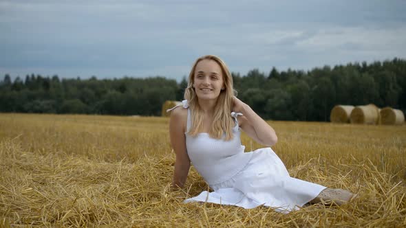 Beautiful Girl in a White Dress Sits and Smiles in a Wheat