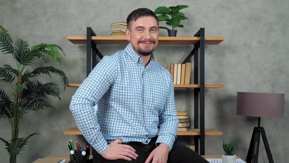 Smiling bearded businessman sits at desk in office turns head looking camera