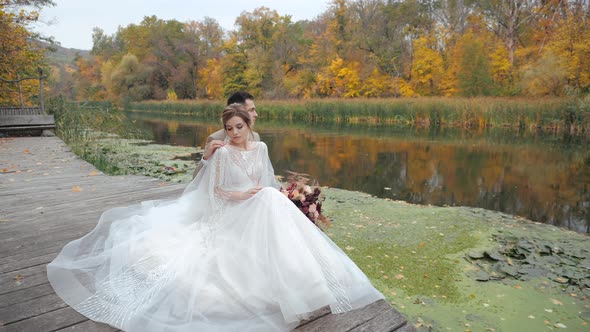 European Young Bride and Groom Are Resting in Nature, Sitting in Embrace on Small Wooden Bridge By