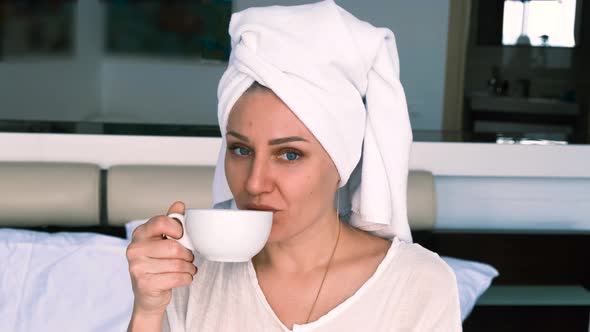 Girl Wearing Bathrobe and Towel Drinks Coffee While Sitting on the Bed Looking at the Camera