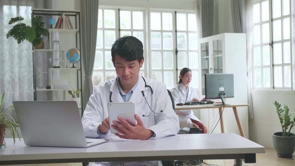 Young Asian Man Doctor Is Using Tablet While Work With Laptop Computer In Workplace