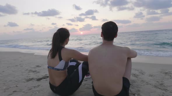 Attractive young couple sit on beach and watch sea waves and beautiful sunset