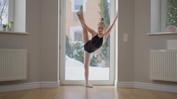 Wide Shot Flexible Talented Teenage Gymnast Stretching One Leg Up Looking at Camera Standing Indoors