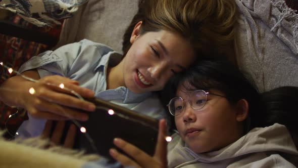 Asian mother and daughter smiling while using digital tablet while lying under blanket fort at home