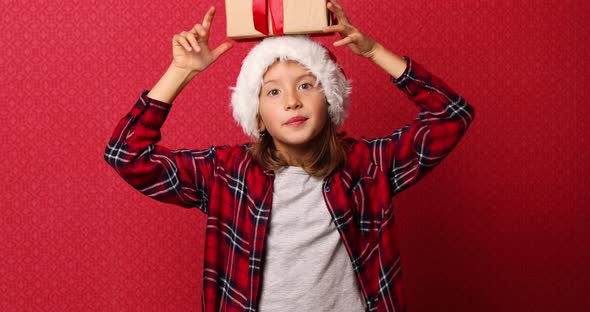 Cute little girl in santa hat with a Christmas gift on her head on isolated red studio background