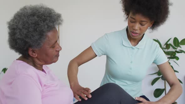 African american female physiotherapist wearing face mask helping senior female patient exercise