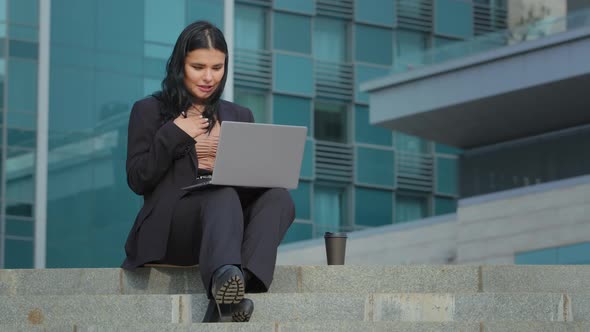 Hispanic Girl Sitting on Steps of Business Center Undergoing Online Interview Using Laptop Presents