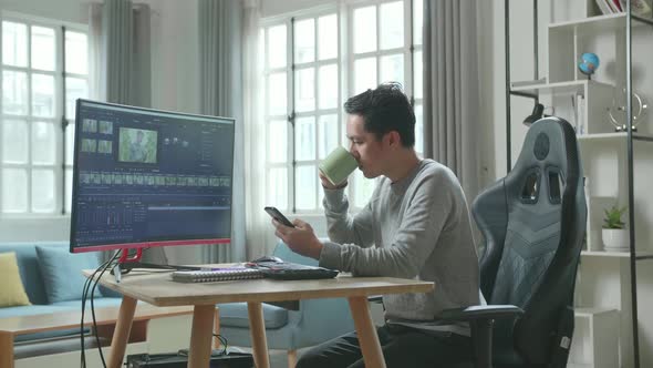 Asian Video Editor Man Using Mobile Phone And Drinking Coffee While Using Computer For Editing Video