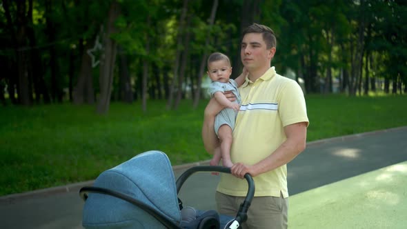 A Young Dad Holds a Newborn Baby in His Arms and Carries a Stroller