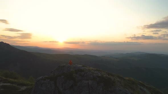 Hiker in Orange Jacket Taking Rest on Rocky Mountain Edge Looking the Sunset