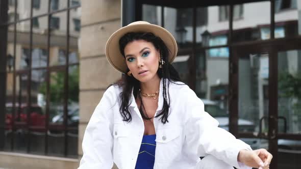 Young Beautiful Brunette in a White Jacket and Brown Hat Sits on the Background of the Building