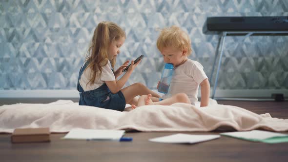 Boy with Bottle Sits on Floor Near Sister with Mobile Phone