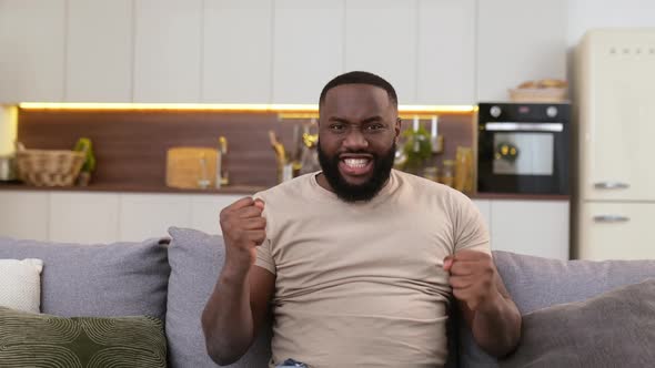 Overjoyed Amazed Excited Young African American Man in Casual Wear Sits in Living Room on a Sofa