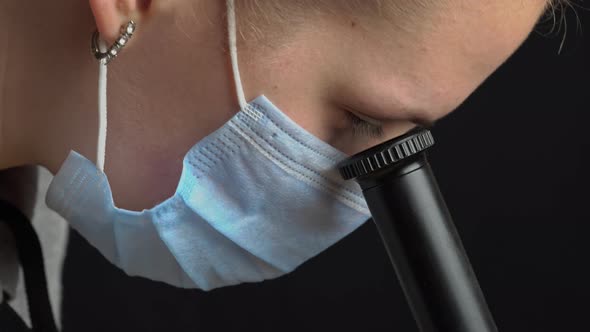 Girl Scientist Looks Into the Lens of a Microscope in a Mask Closeup