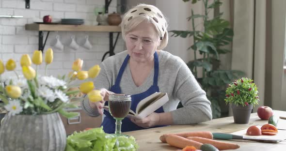 Portrait of Calm Caucasian Senior Woman Reading Book and Drinking Red Wine in Kitchen. Fresh