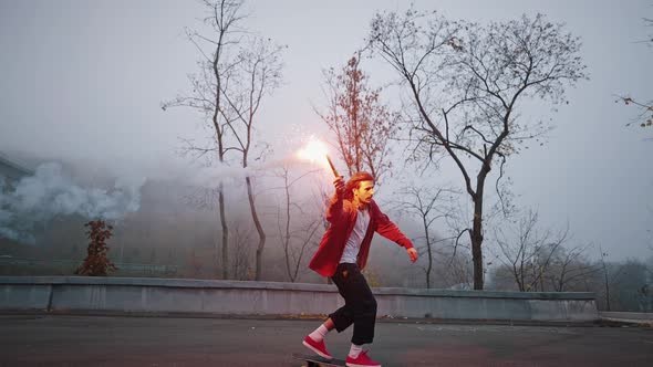 Young Male Skater Holds Colored Red Smoke Bomb in His Hand While Riding a Longboard in the City Park