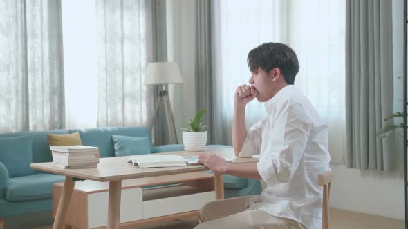 Side View Of Asian Man Student Being Sleepy While Writing In The Notebook On The Table At Home