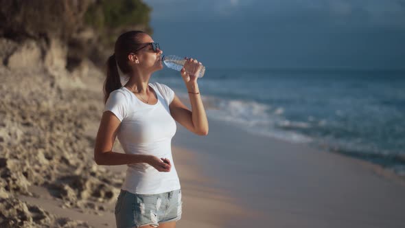 Beautiful Young Woman in Sunglasses Drinks Clear Water From Bottle on Beach