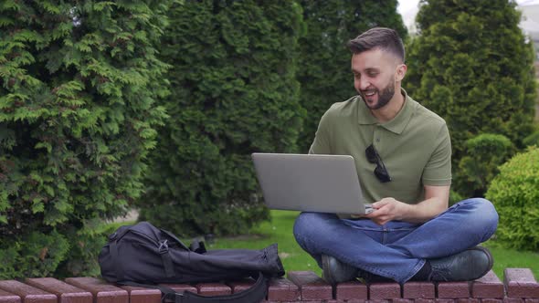Young Man with Laptop Sitting on a Wooden Bench in Park