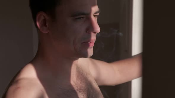 Close-up young muscular man smoking a cigarette and looks out the window