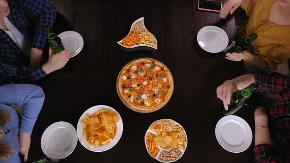 A Group of Happy Friends Clink Glasses of Beer Over a Table with Pizza and Chips