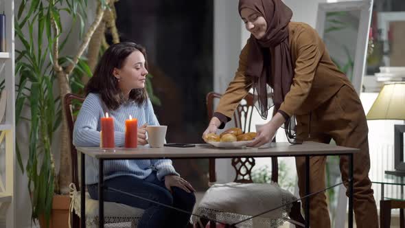 Young Caucasian Woman Drinking Tea As Middle Eastern Friend Bringing Homemade Buns