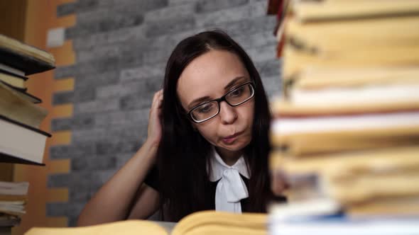 Tired Female Student Reading Among Books. Pensive Young Woman Sitting at Table with Pile of Book and