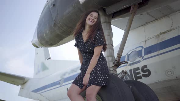 Happy Young Brunette Woman in Black Dress Looking at the Camera Sitting on the Landing Gear